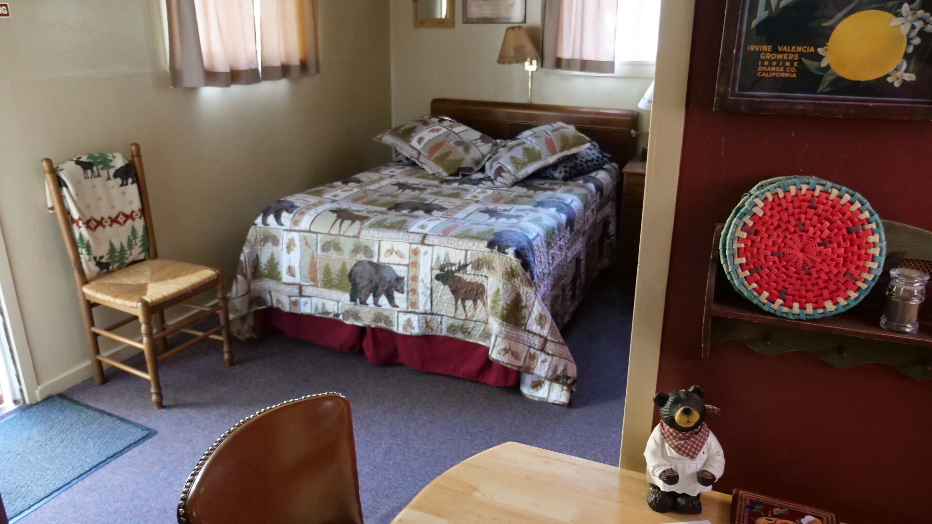 A cozy bedroom with a bed covered in a wildlife-themed quilt, a wooden chair, a small table, and a decorative bear figurine.