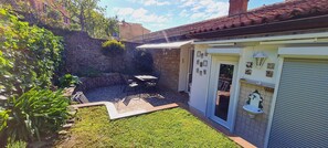 Un pequeño patio con muro de piedra, una mesa y sillas, y una puerta blanca con entrada de azulejos.