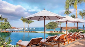Un área junto a la piscina con sillones reclinables y sombrillas, palmeras y vista al océano.