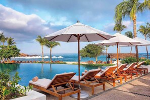 Ein Poolbereich mit Liegestühlen und Sonnenschirmen, Palmen und Blick auf das Meer.