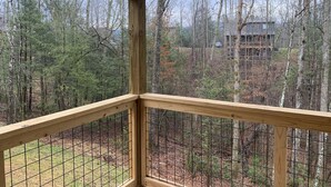 A wooden deck with a grid railing overlooking a forested area and a house in the background. - Cozy Cabin With Fire Pit and Private Hot Tub (Gatlinburg)
