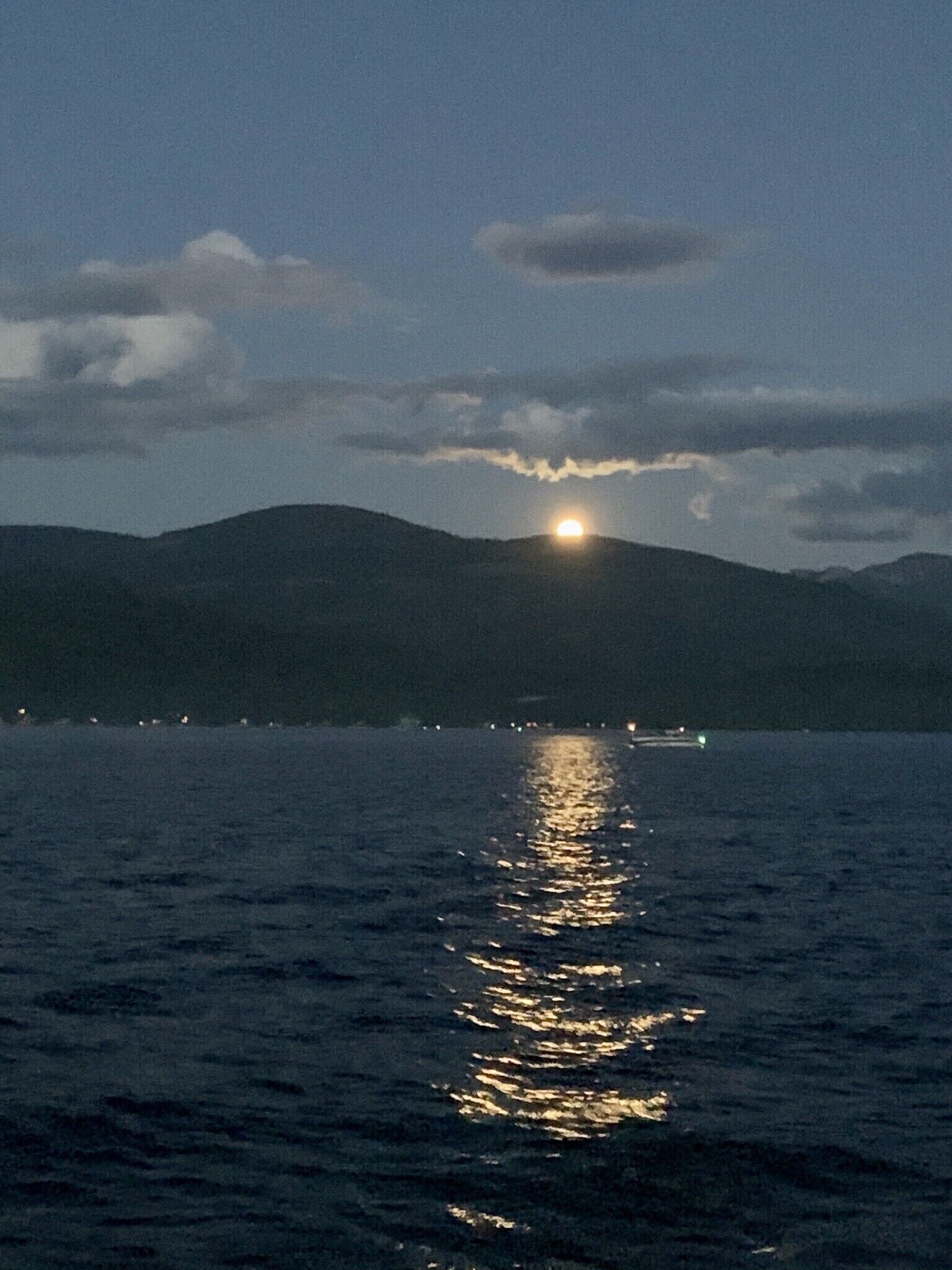 Une vue nocturne d’une étendue d’eau reflétant la lune et les lumières lointaines, avec des montagnes en arrière-plan.