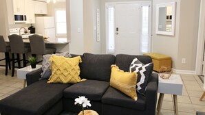 A modern living room with a dark grey sectional sofa, a small white side table, and a yellow ottoman. A kitchen with white cabinets and a dining area with dark chairs are visible in the background.