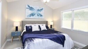 A bedroom with a bed, two bedside tables, a ceiling fan, a window with blinds, and a wall-mounted abstract painting.