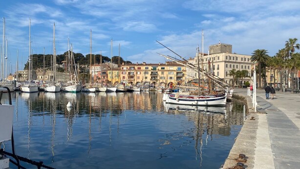 Un puerto deportivo con barcos amarrados, un paseo marítimo y edificios al fondo.