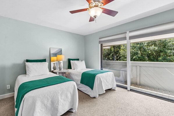 A hotel room with two beds, a ceiling fan, a large window, and a view of greenery outside.