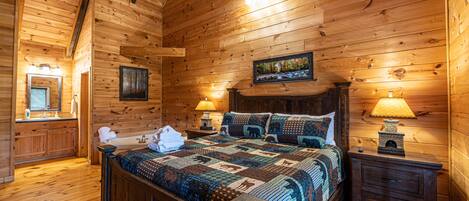 A wooden cabin bedroom with a large bed, two bedside lamps, a nightstand, and a framed picture on the wall.
