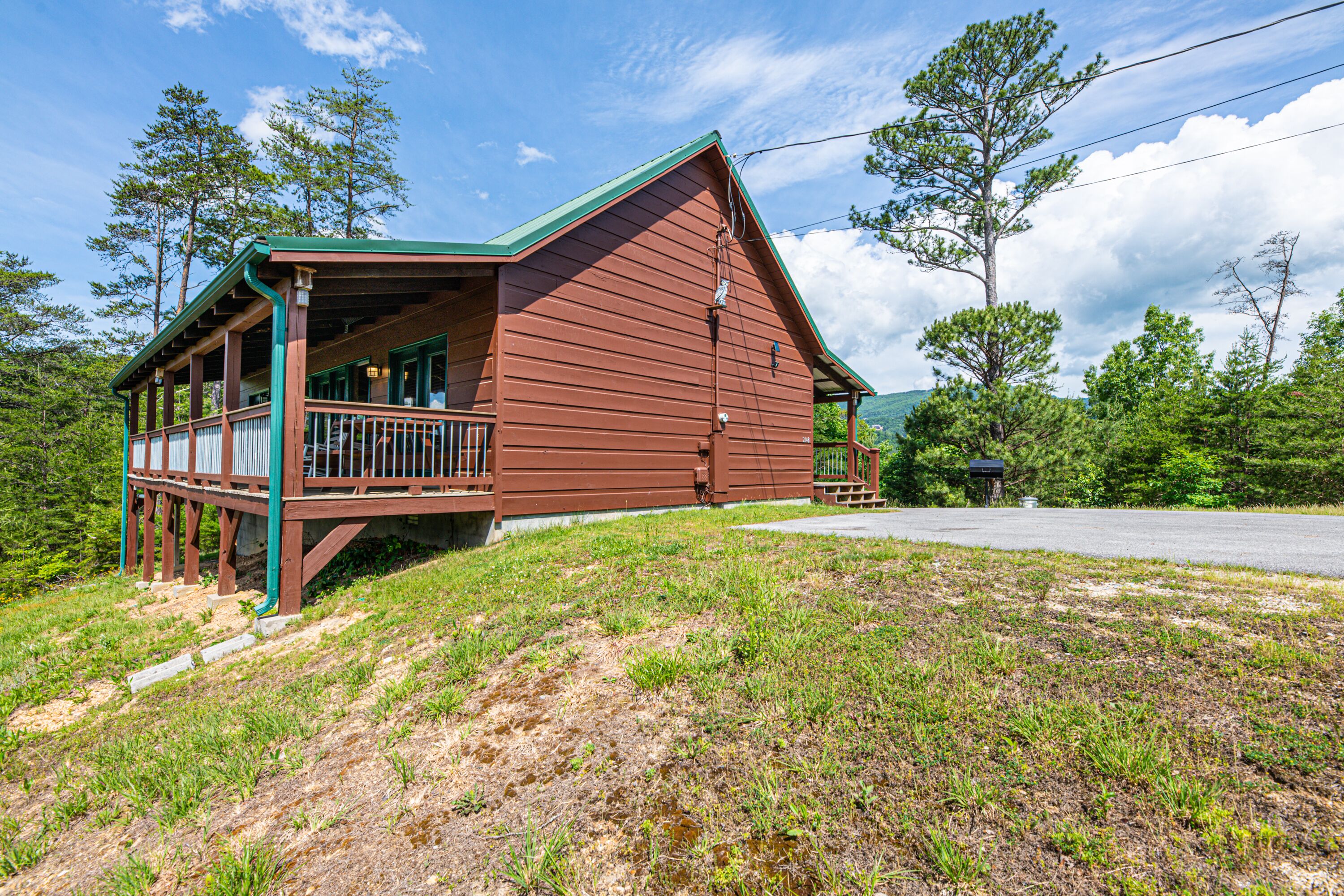 A wooden cabin with a porch and a covered deck, surrounded by trees and a grassy area.