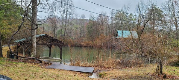 A rustic wooden gazebo by a calm lake, surrounded by dense trees and a distant house. - Stunning View Retreat: 3 Rooms, Hot Tub, Fire Pit & 10 Minutes to Pigeon Forge (Sevierville)