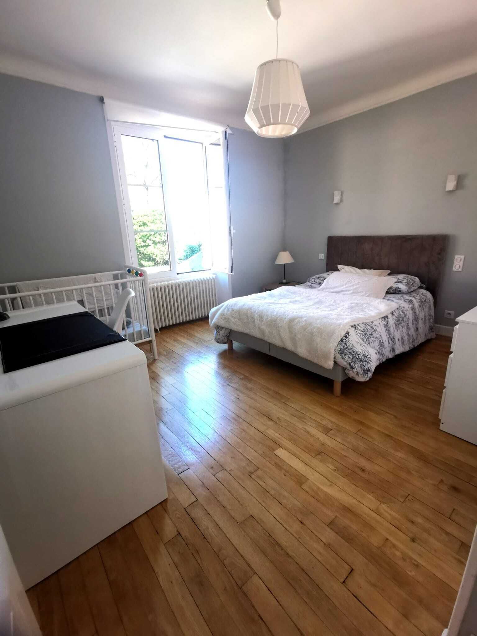 A bedroom with a bed, a white chair, a radiator, a window, and a hanging light fixture.