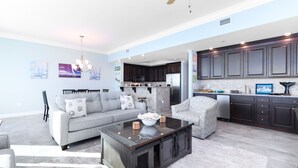 A spacious living room with a gray sofa, a coffee table, and a patterned armchair. A kitchen with dark cabinets and a refrigerator is visible in the background. - Phoenix Orange Beach II 1003 by Liquid Life (Orange Beach)