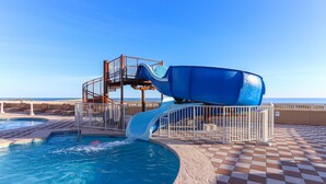 Uma piscina com escorregador azul, um deck ensolarado com padrão xadrez e vista clara do oceano.