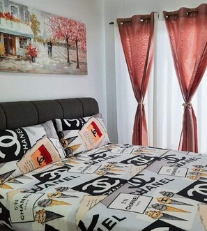 Quarto com cama coberta por edredom com tema Chanel, um quadro na parede e cortinas vermelhas.