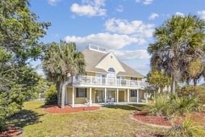 Stunning two-story home with a central entrance and peaked roof, set amidst a lush landscape of palm and pine trees.