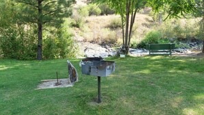 Uma churrasqueira em uma área gramada, com um banco e árvores ao fundo.