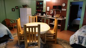 Quarto acolhedor com mesa de jantar de madeira rodeada por cadeiras, pequena cozinha e cama com manta estampada.