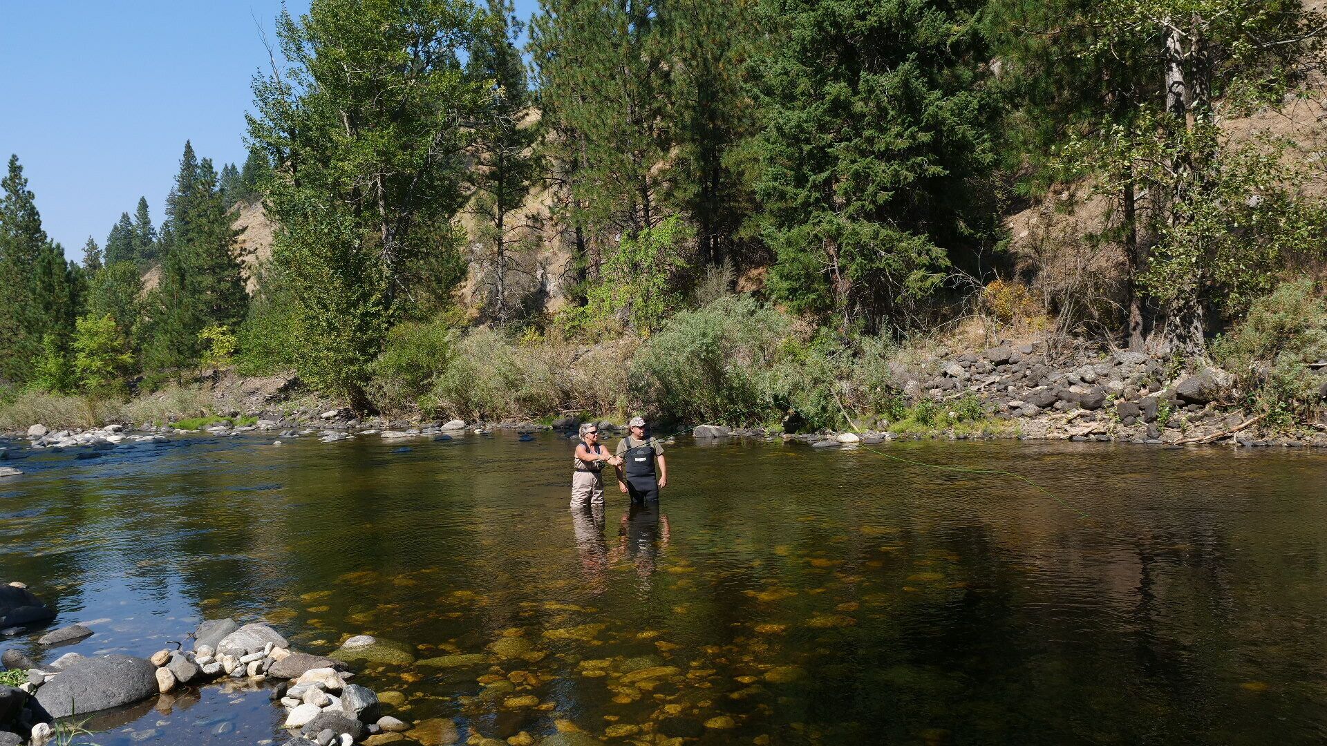 A river with clear water, surrounded by dense forest and rocky terrain.