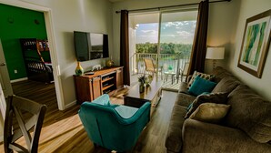 Ein Wohnzimmer mit Sofa, einem türkisfarbenen Sessel, einem Fernseher, einer Holzkabine und einem Balkon mit Blick ins Grüne.