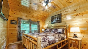 A wooden cabin bedroom with a bed, a nightstand, a ceiling fan, and a window with curtains.