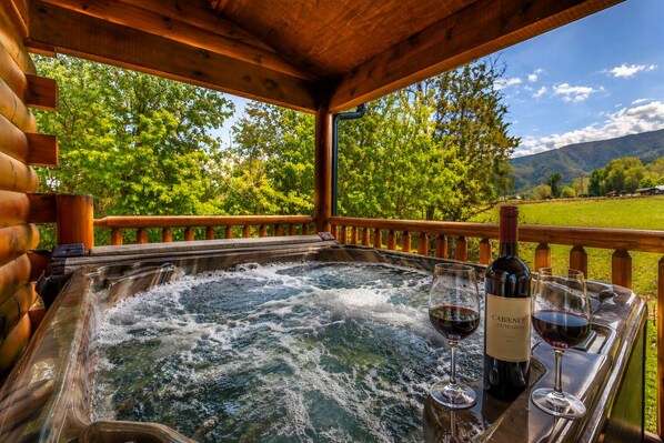 A hot tub with a wooden deck, surrounded by lush greenery and a mountainous backdrop.