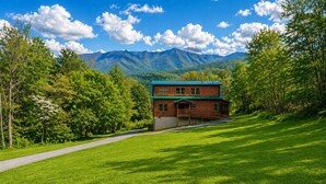 A two-story log cabin with a green roof, surrounded by lush green grass and trees, with mountains in the background.