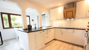 A modern kitchen with white cabinetry, a black countertop, and a dishwasher. There is a window with a view of greenery outside. - Weiße Höhen - Ferienhaus, Dibbersen bei Hamburg (Buchholz in der Nordheide)