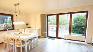 A modern dining area with a white table and chairs, a pendant light, and a view of a garden through large sliding glass doors. - Weiße Höhen - Ferienhaus, Dibbersen bei Hamburg (Buchholz in der Nordheide)