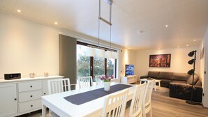 A modern living room with a white sofa, a dining table with chairs, and a kitchen area with cabinets and a countertop. - Weiße Höhen - Ferienhaus, Dibbersen bei Hamburg (Buchholz in der Nordheide)