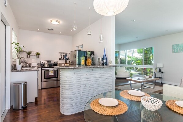A modern kitchen with a central island, stainless steel appliances, and a dining area with glass tables and woven placemats. - Tropical Paradise Beach Getaway, Villa #1 (Lauderdale-by-the-Sea)
