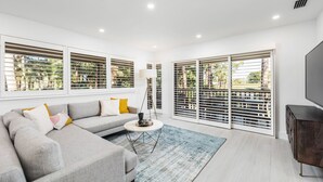 Una sala moderna con un sofá seccional gris, una mesita redonda pequeña y un televisor sobre un mueble de madera. Amplias ventanas con persianas ofrecen vistas al verdor exterior.