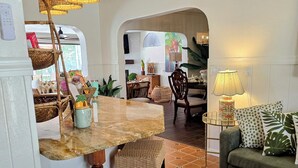 A cozy living room with a green sofa, wicker chairs, a dining area with a marble table, and a kitchen visible through an archway. - Enchanted Private Island Oasis Steps To Beach, Tropical Yard & Charm Filled Home (Placida)