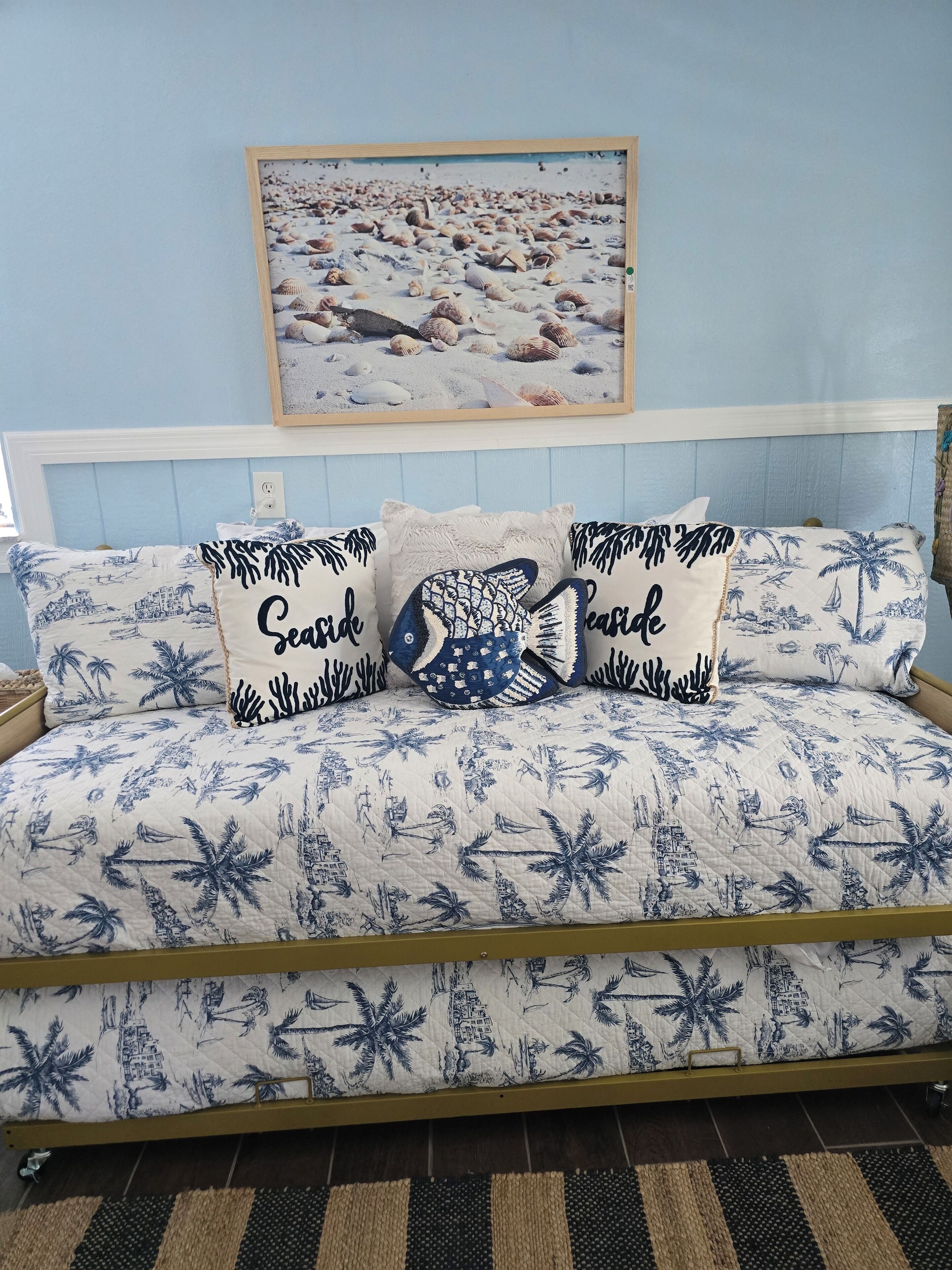 A bed with a blue and white tropical print bedspread, a framed beach scene above it, and a matching pillow with the word "peaceful" on it.