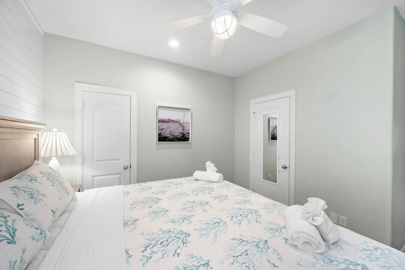 A bedroom with a bed, a ceiling fan, a nightstand with a lamp, and a framed picture on the wall.
