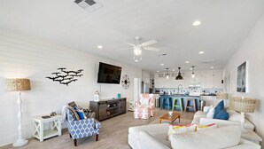 Un salon spacieux avec un canapé blanc, un fauteuil à motifs et une table basse. Un téléviseur est fixé au mur, et on aperçoit un coin cuisine au fond de la pièce.