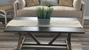 Een woonkamer met een beige bank, twee gekleurde kussens, een kleine potplant en een donker houten salontafel met een geweven onderzetter.