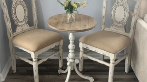 Twee antieke, crèmekleurige stoelen met gewatteerde zittingen en rugleuningen, een klein rond houten tafeltje met een bloevas, en een witte, versierde spiegel aan de muur.