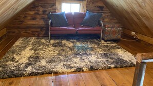 Une chambre mansardée avec un canapé rouge, deux coussins, une télévision et un tapis à motifs.