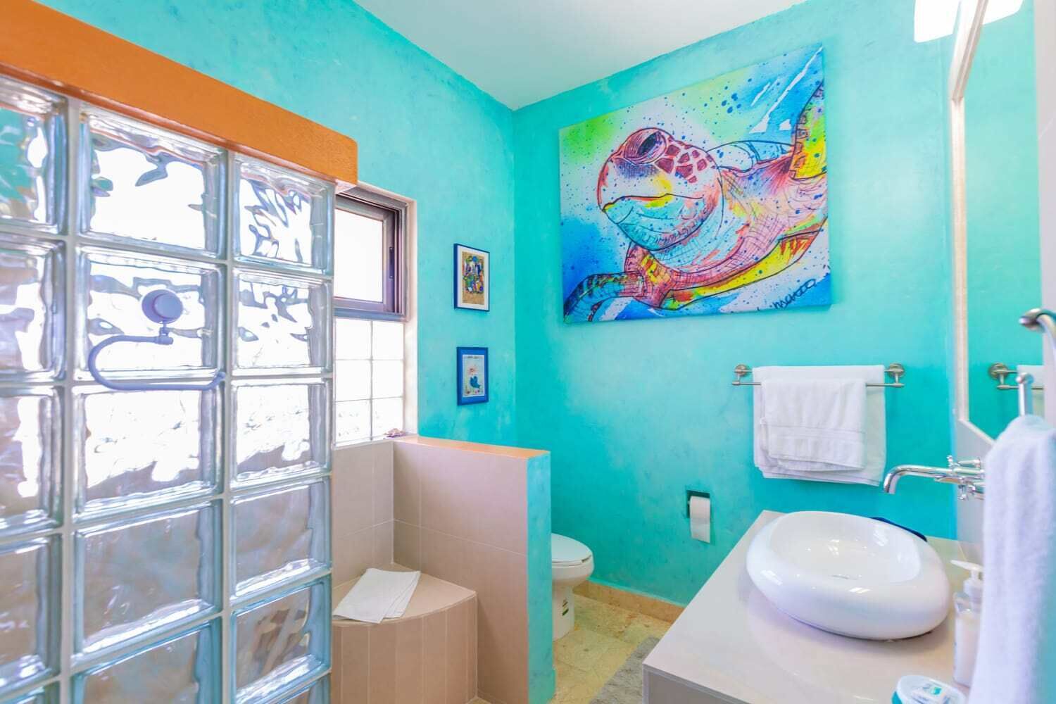 A bathroom with a turquoise wall, a glass shower enclosure, a white sink, and a colorful painting.