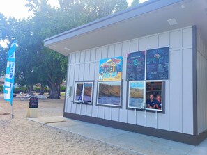 Un petit kiosque de restauration situé sur la plage, avec un panneau affichant le menu, une pancarte indiquant « Retrait des commandes ici » et une fenêtre pour passer les commandes.