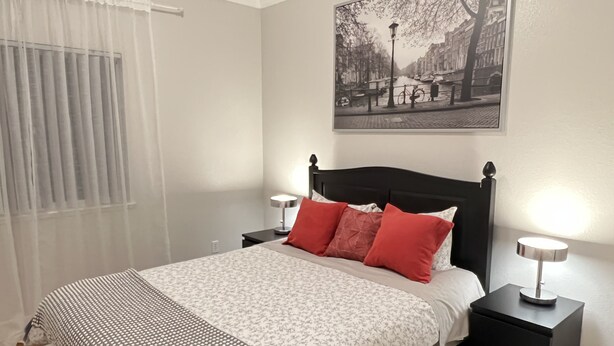 Una camera da letto ordinata con un letto, due comodini, una finestra con tende leggere e un dipinto con vista sulla città.