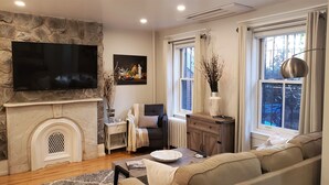 A living room with a stone fireplace, a flat-screen TV mounted on the wall, a sofa, a coffee table, and a window with curtains. - Modern, Smart 1Bed -Private Entry. Boston's Best Location Close to Everything (Boston)