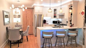 A modern kitchen with white cabinets, a stainless steel refrigerator, a built-in island with bar stools, pendant lighting, and a wooden door. - Modern, Smart 1Bed -Private Entry. Boston's Best Location Close to Everything (Boston)