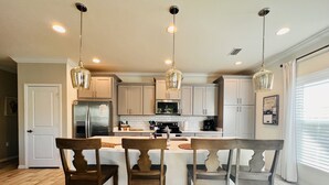 Una cocina moderna con armarios blancos, una isla central con taburetes de bar, electrodomésticos de acero inoxidable e iluminación colgante.