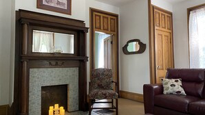 A living room with a fireplace, a floral armchair, a leather sofa, and a mirror on the wall. - Hillside Haven Historic Vacation Home in Deersville by Tappan & Clendening Lakes (Deersville)