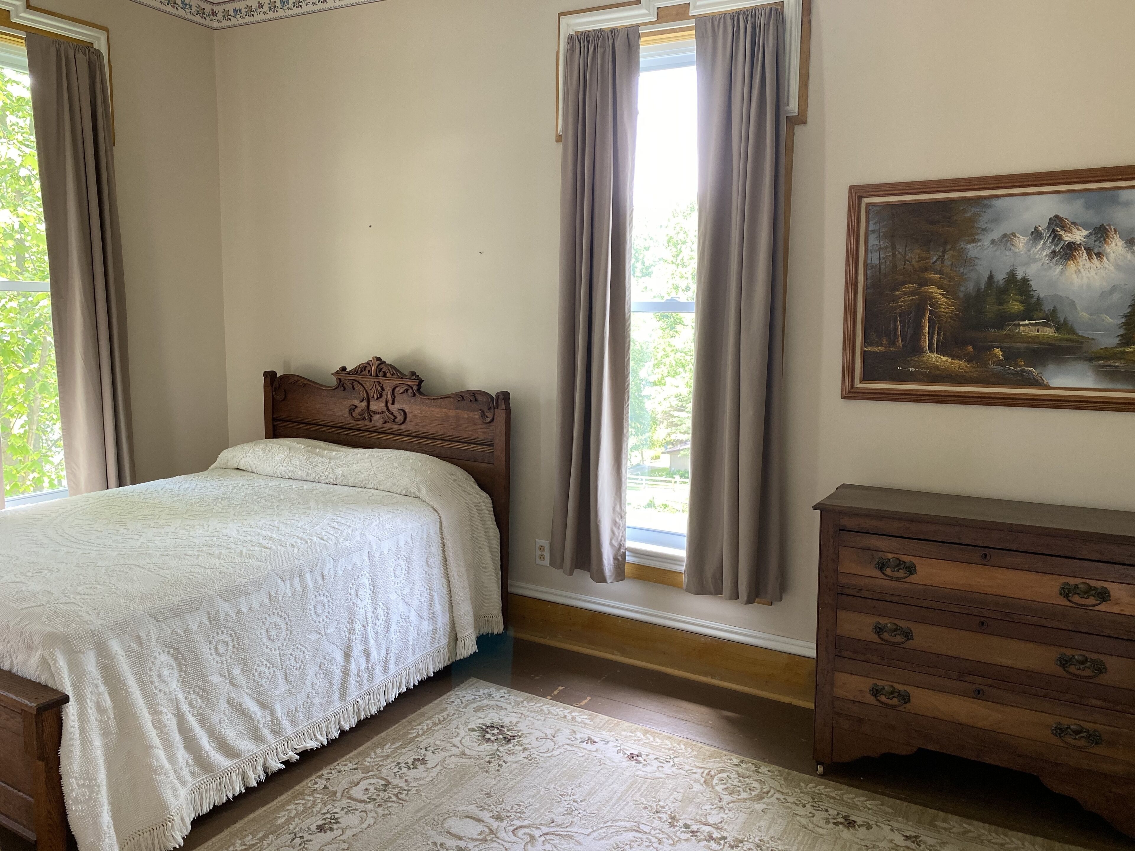 Une chambre à coucher avec un lit en bois, une commode, un tableau et une vue sur la verdure par la fenêtre.