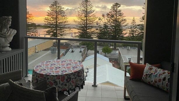 A balcony with a view of a sunset, a round table with a floral tablecloth, and a sofa with patterned cushions. - Oceanview Escape at The Entrance (The Entrance)