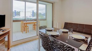 A dining area with a table set for four, a television on a stand, and a view of a cityscape through large windows. - Horizon Océan 1 by Interhome (Capbreton)