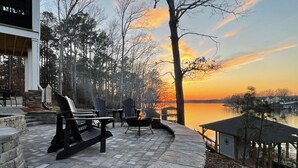 Un espace patio aménagé avec des fauteuils Adirondack, un foyer et une vue sur un lac au coucher du soleil.