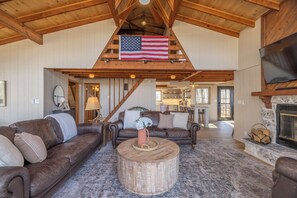 A spacious living room with a high ceiling, a large American flag, leather sofas, a round coffee table, and a fireplace.