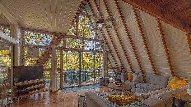 An attic-style room with a large TV, wooden furniture, and a view of trees through large windows. - Rae of Sunshine by Innsbrook Vacations! (Innsbrook)